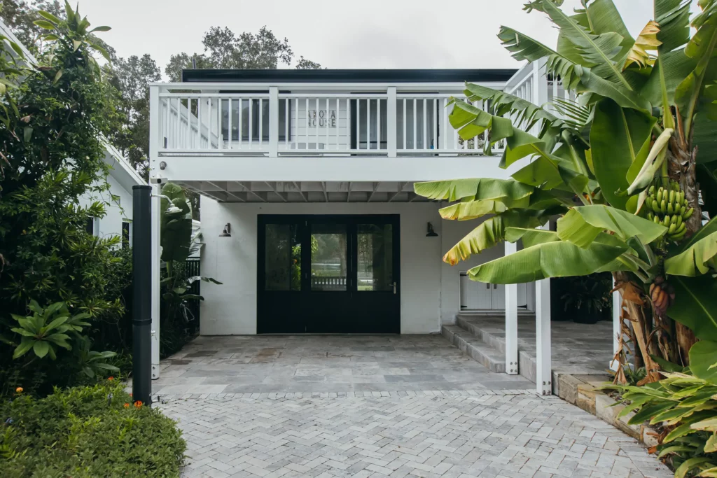 Landscape photo of exterior of home. House is two stories with a carport and a balcony above. The home has a coastal feel with white timber and a lush green garden.